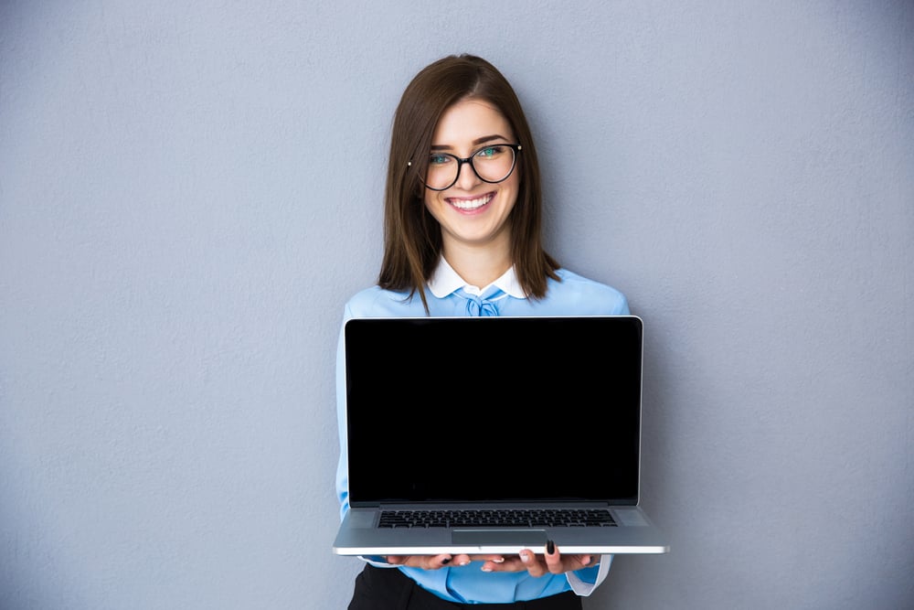 Happy businesswoman showing blank laptop screen over gray background. Wearing in blue shirt and glasses. Looking at camera Happy businesswoman showing blank laptop screen over gray background. Wearing in blue shirt and glasses. Looking at camera