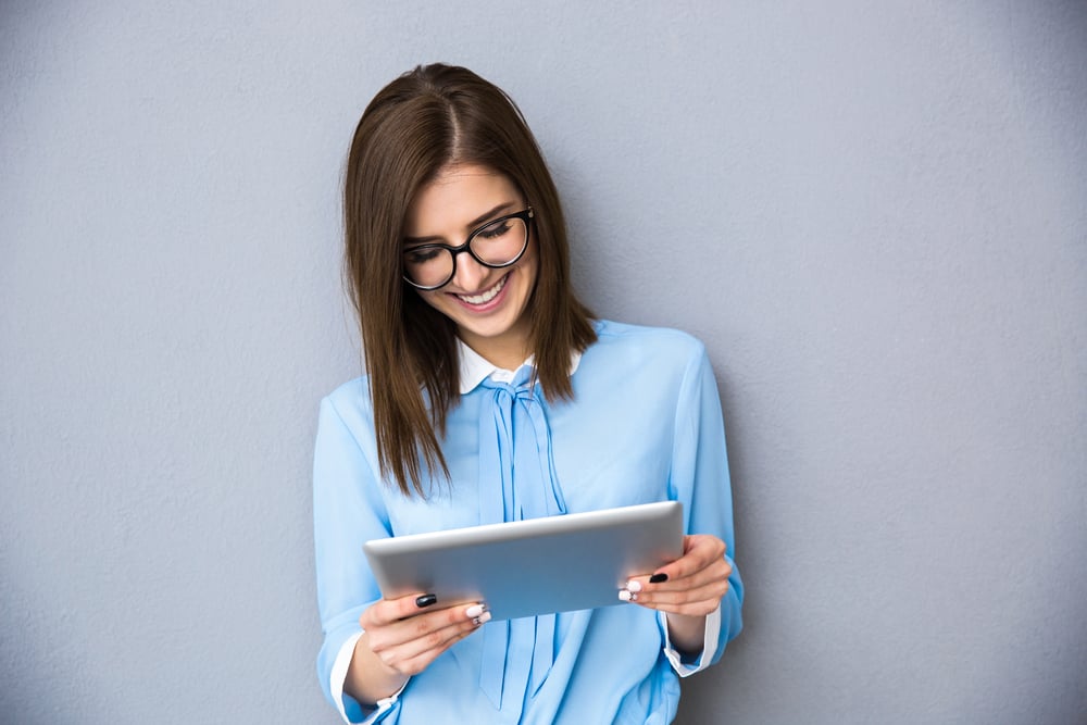 Happy businesswoman standing with table computer over gray background. Wearing in blue shirt and glasses. Happy businesswoman standing with table computer over gray background. Wearing in blue shirt and glasses.